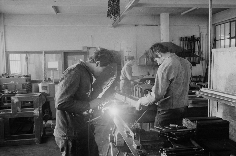 Drei Lehrlinge arbeiten in einer Schlosserwerkstatt. Im Vordergrund wird geschweisst, weiter hinten Metall mit Werkzeugen bearbeitet.