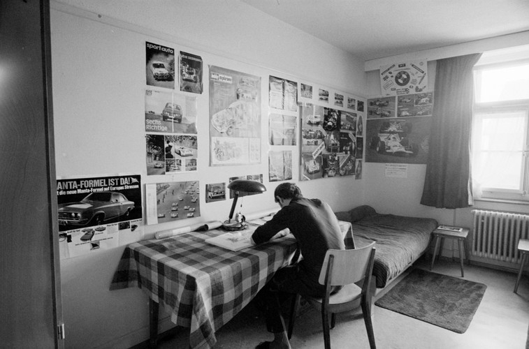 Schwarz-Weiss Foto eines Zimmers in der Erziehungsanstalt Uitikon-Waldegg. Ein Jugendliche sitzt an seinem Arbeitstisch, links davon steht ein Bett mit einem Teppich davor. Am Fenster hängen Gardinen und an den Wänden Poster von Sportwagen.