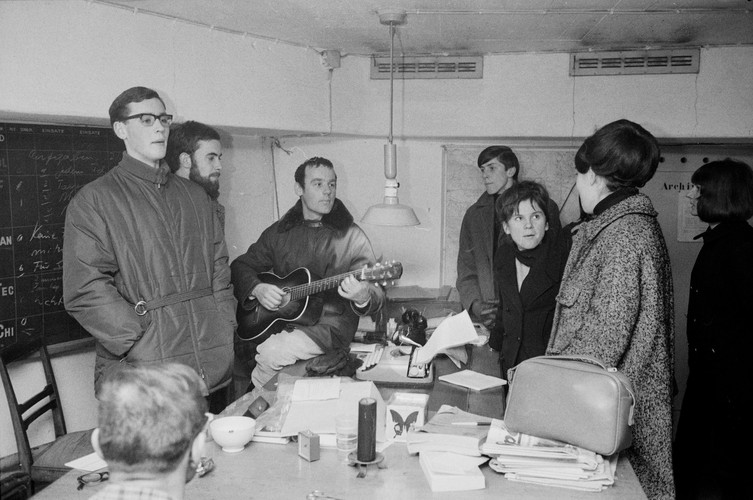 Schwarz-Weiss-Foto mit dem jungen Pfarrer Ernst Sieber in der Mitte, eine Gitarre haltend, in einem Bunkerraum. Um einen Tisch herum stehen oder sitzen sechs weitere Personen, alle in winterlicher Kleidung. Sie hören der Musik zu oder unterhalten sich.