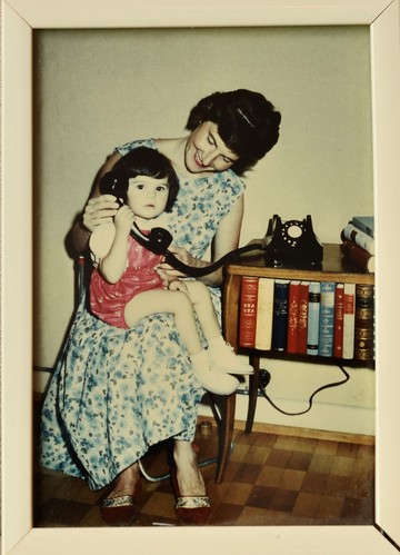 Die circa 2-jährige Jasmin Schweizer* sitzt auf dem Schoss ihrer Mutter und hält einen grossen schwarzen Telefonhörer in der Hand. Ihre Mutter beugt sich zu ihr und lächelt. Ein schwarzes Telefon mit weisser Wählscheibe steht auf einem kleinen Büchergestell rechts daneben.